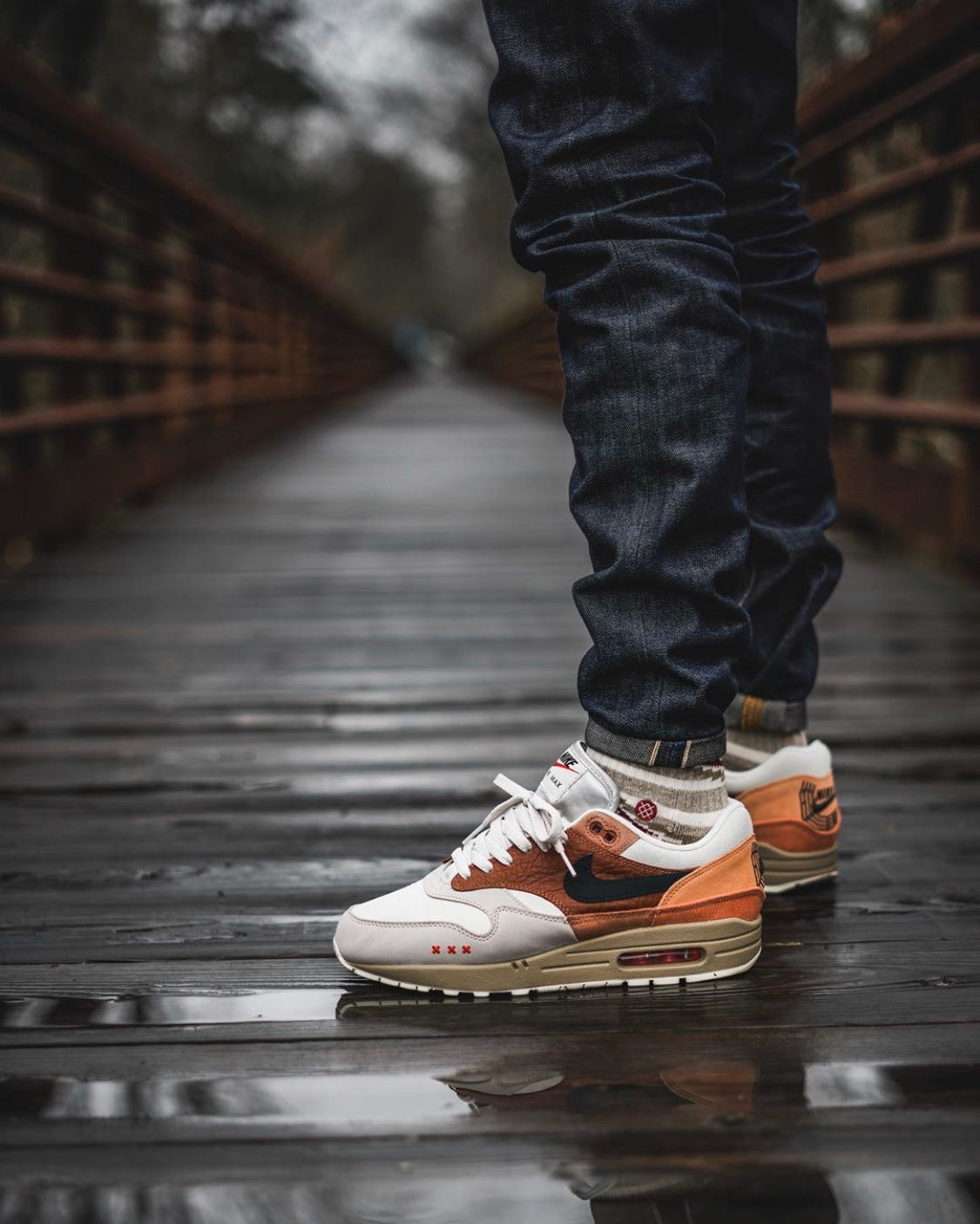 Person wearing Nike Air Max 1 Amsterdam City Pack sneakers in white, brown, and orange standing on a wet wooden bridge, showcasing exclusive European street style.