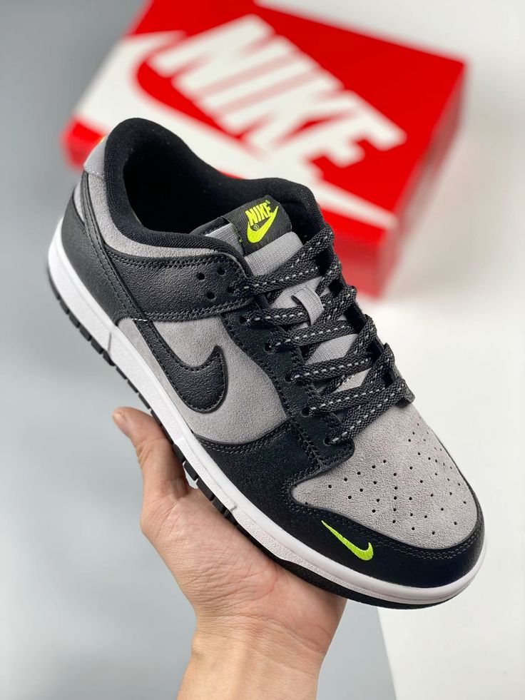 Hand holding a Nike Dunk Low Black Grey Green Strike sneaker featuring black and grey suede with neon green accents, against a blurred red and white Nike box background, showcasing exclusive sneakers available across Europe. The real deal.