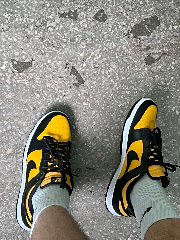Person wearing Nike Dunk Low Black University Gold sneakers with black and gold color accents and white soles, standing on a textured gray surface. The real deal.