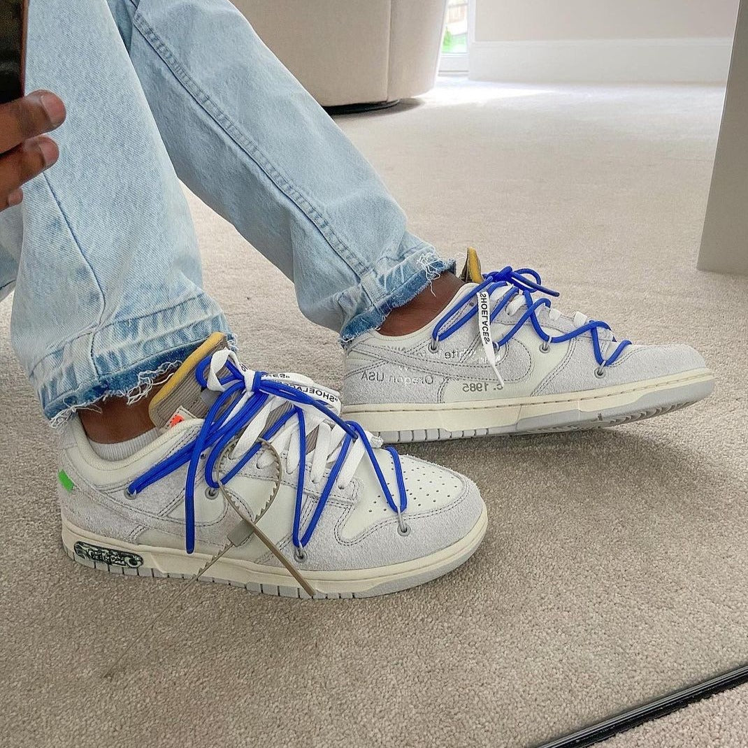 Person wearing Nike Dunk Low Off-White Lot 32 sneakers with bright blue laces and light wash jeans, sitting indoors on carpeted floor, showcasing exclusive European sneaker style The real deal