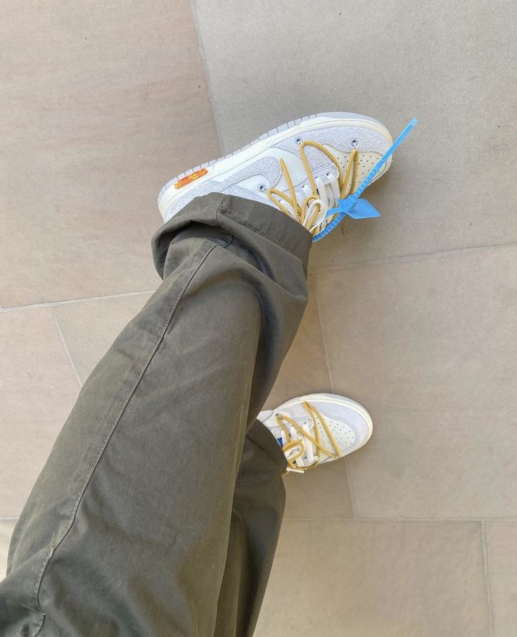 Person wearing white and beige Nike Dunk Low Off-White Lot 34 sneakers with yellow laces and a blue tag, paired with olive green pants on a tiled floor, showcasing exclusive sneaker style from whole Europe and The real deal.