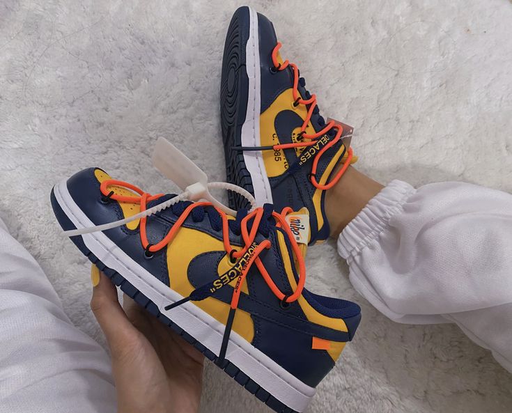 A pair of Nike Dunk Low Off-White Michigan sneakers in navy blue and yellow with bright orange laces, styled on feet with white pants, showcasing exclusive European streetwear style.