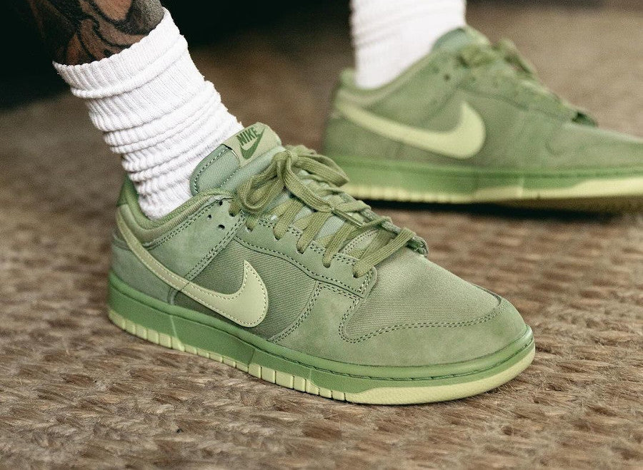 Close-up of Nike Dunk Low Premium Oil Green sneakers worn with white ribbed socks, featuring premium suede panels in varying shades of green and a classic Nike swoosh, showcasing exclusive European sneaker style. The real deal.