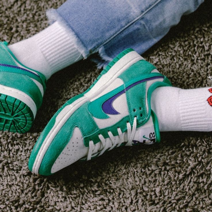 Person wearing Nike Dunk Low SE 85 Neptune Green sneakers with white socks and blue jeans, sitting on a textured carpet, highlighting the exclusive design and vibrant colors of the shoes, the real deal.