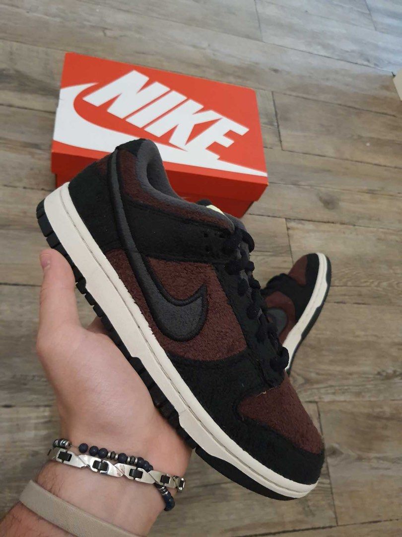 Hand holding a Nike Dunk Low SE Fleece Burgundy Crush sneaker in front of a red Nike shoebox on a wooden floor, showcasing exclusive European sneakers - The real deal.