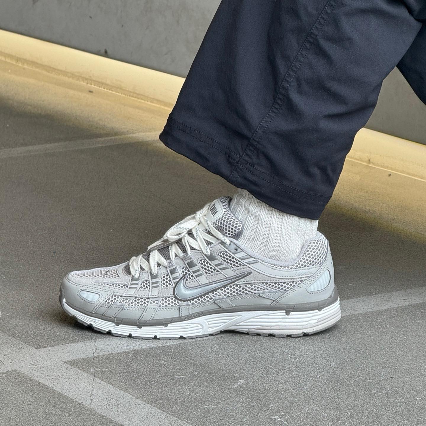 Person wearing white socks and Nike P-6000 Premium Light Iron Ore sneakers with grey and silver detailing, standing on a pavement surface, showcasing exclusive European sneaker style.