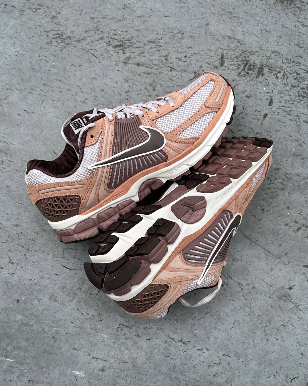 Pair of Zoom Vomero 5 Dusted Clay sneakers in light brown and white mesh with dark brown accents, displayed on a concrete surface highlighting their design and sole tread.