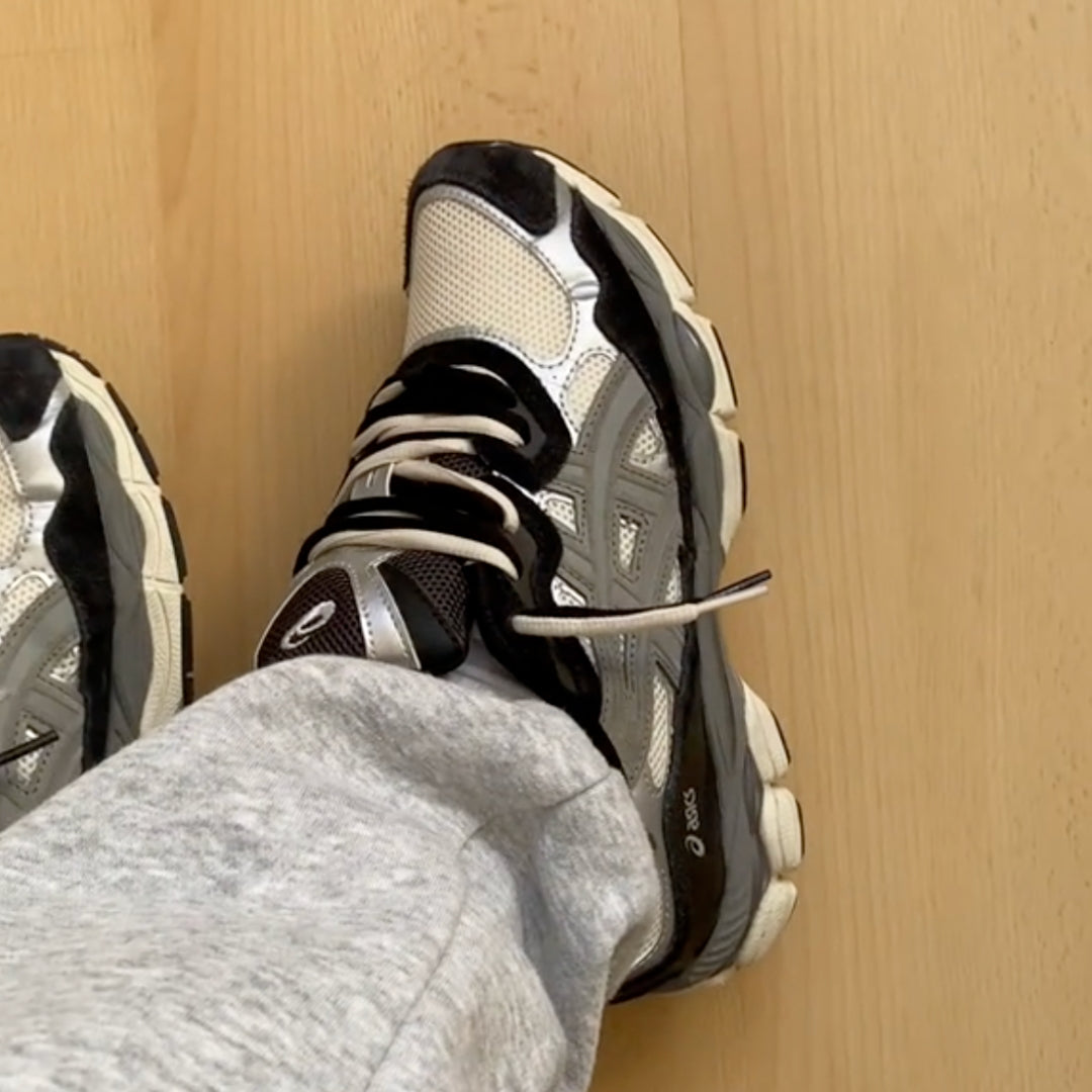 Person wearing ASICS Gel-NYC Ivory Clay Grey sneakers paired with grey sweatpants on a light wooden floor, showcasing exclusive trainers in Europe branded as The real deal.