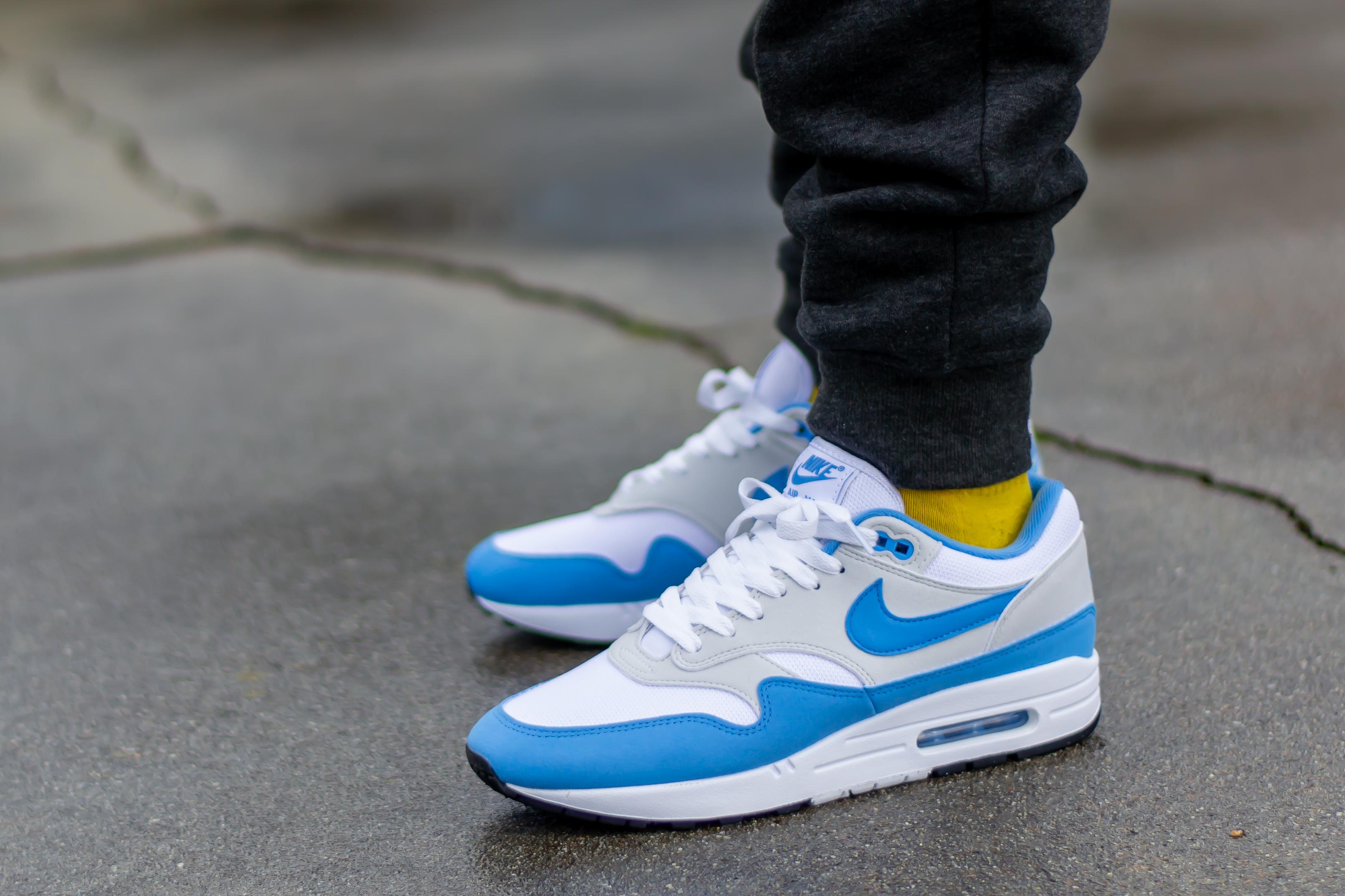 Person wearing Nike Air Max 1 White University Blue sneakers with yellow socks and black pants standing on a concrete surface, showcasing exclusive European sneaker style.